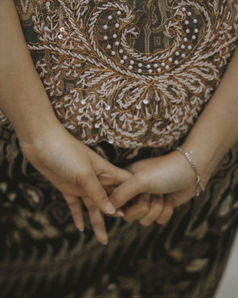 Hands with ivory tipped nails folded over gold embroidered fabric