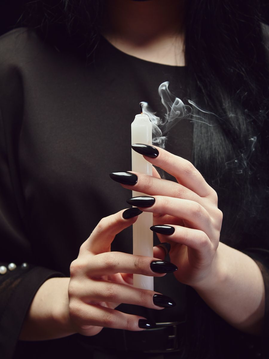 A hand with black-polished nails holding a lit taper candle in a dark room.