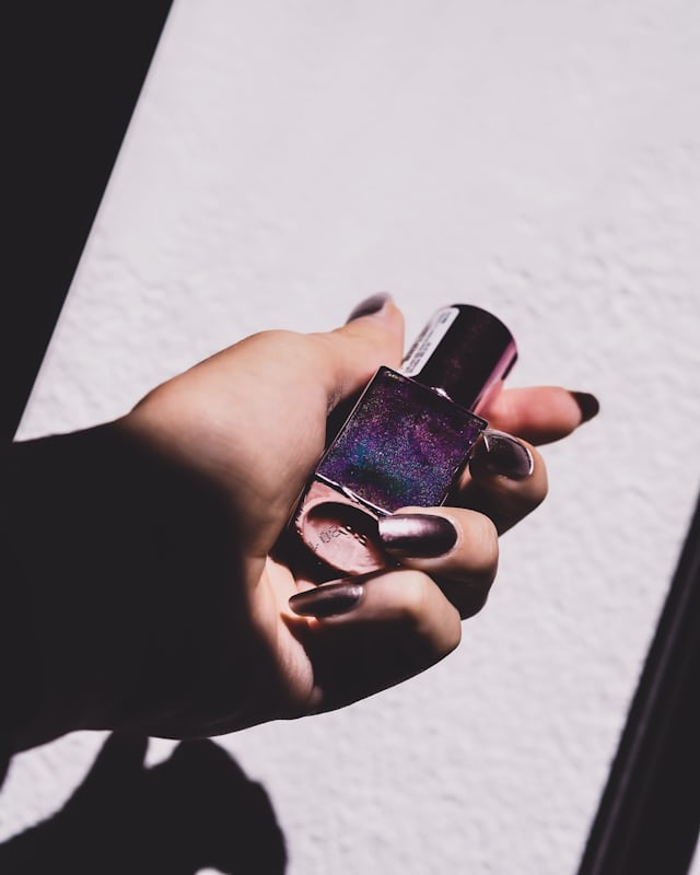 A hand holding a bottle of deep purple polish in hard sunlight